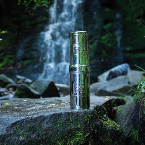 An American Reserves British Berkefeld 1 Liter Stainless Steel Gravity System & Ultra Sterasyl sits on a mossy rock, gleaming as ultra-pure water flows from the waterfall, surrounded by lush greenery.