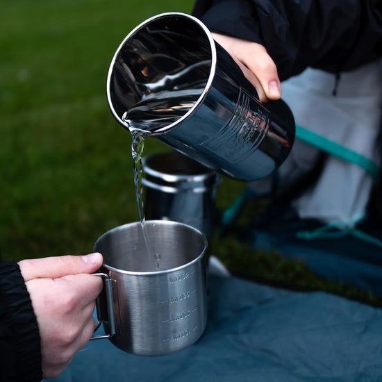 Two hands hold metal cups amidst grass, one pouring liquid into the other, showcasing the efficiency of an American Reserves British Berkefeld 1 Liter Stainless Steel Gravity System & Ultra Sterasyl.