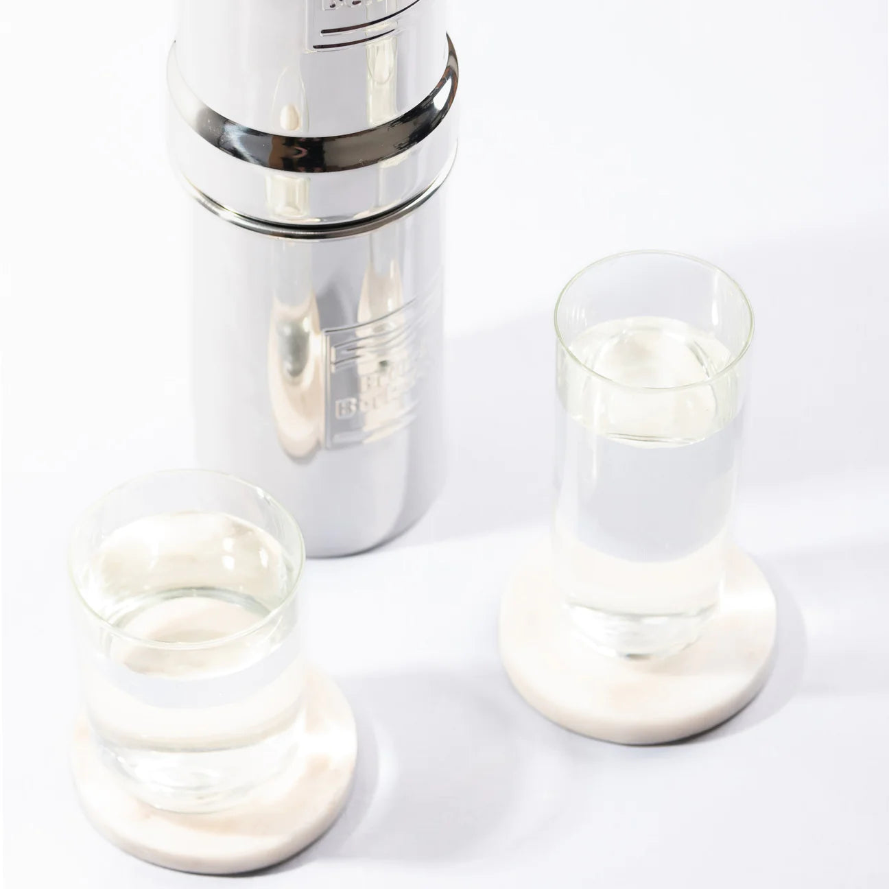 The British Berkefeld 1 Liter Stainless Steel Gravity System & Ultra Sterasyl by American Reserves, placed on a white surface with two glasses of water on coasters, exemplifies efficiency and elegance.