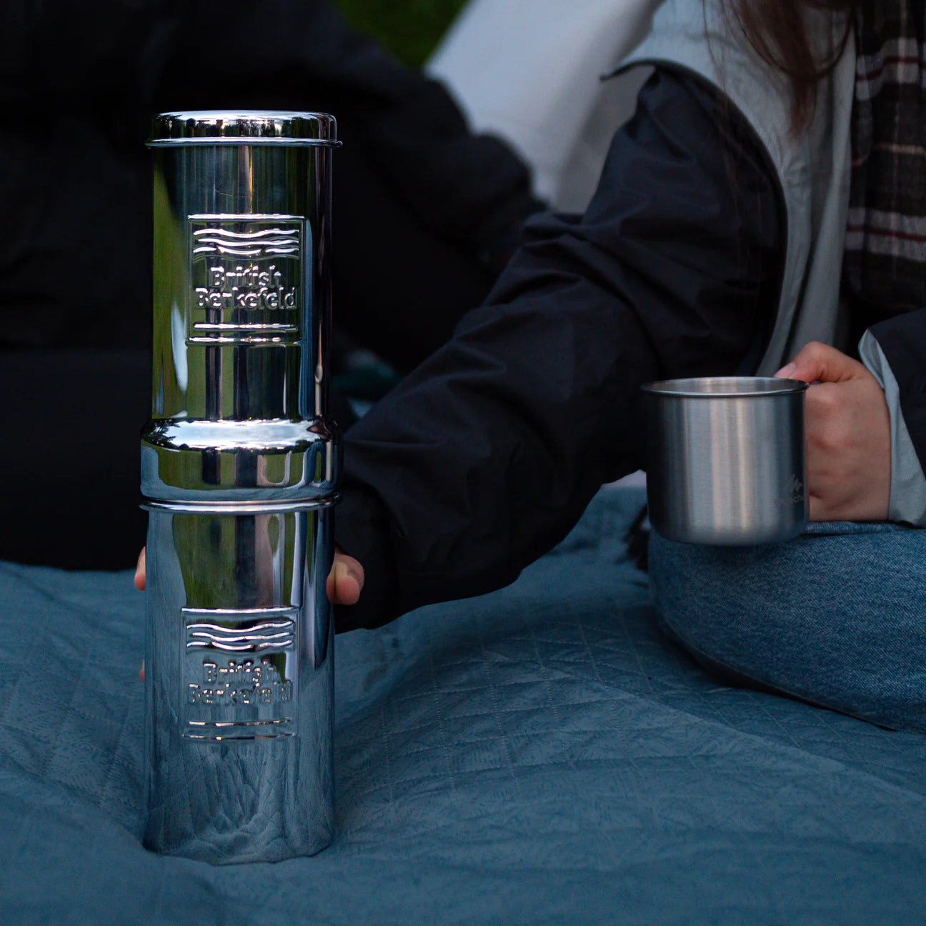 A person sits on a blanket, holding a metal cup beside a tall stack of metal containers. The standout is the British Berkefeld 1 Liter Stainless Steel Gravity System by American Reserves, featuring an Ultra Sterasyl ceramic filter for optimal water purity.