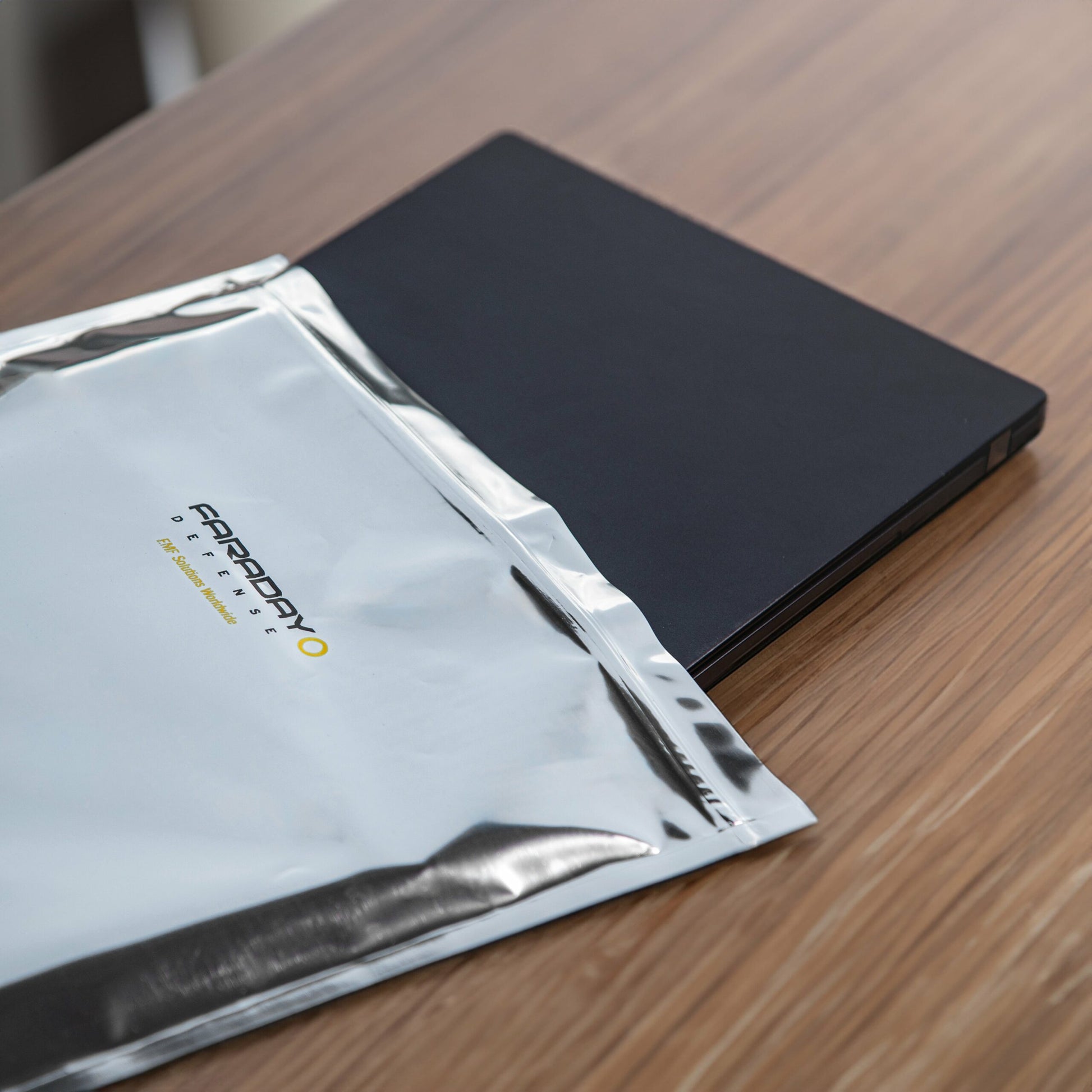 A black laptop is partially placed inside a silver zip-lock closure bag from the 15pc 7.0 Mil Faraday EMP Kit by American Reserves on a wooden surface, ensuring strong electromagnetic radiation protection.