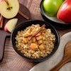 A bowl of oatmeal with chopped apples and cinnamon sits on a wooden table, with American Reserves 30 Day Emergency Food Supplys freeze-dried apples nearby, alongside whole apples and a cloth napkin—ideal for long-term storage.