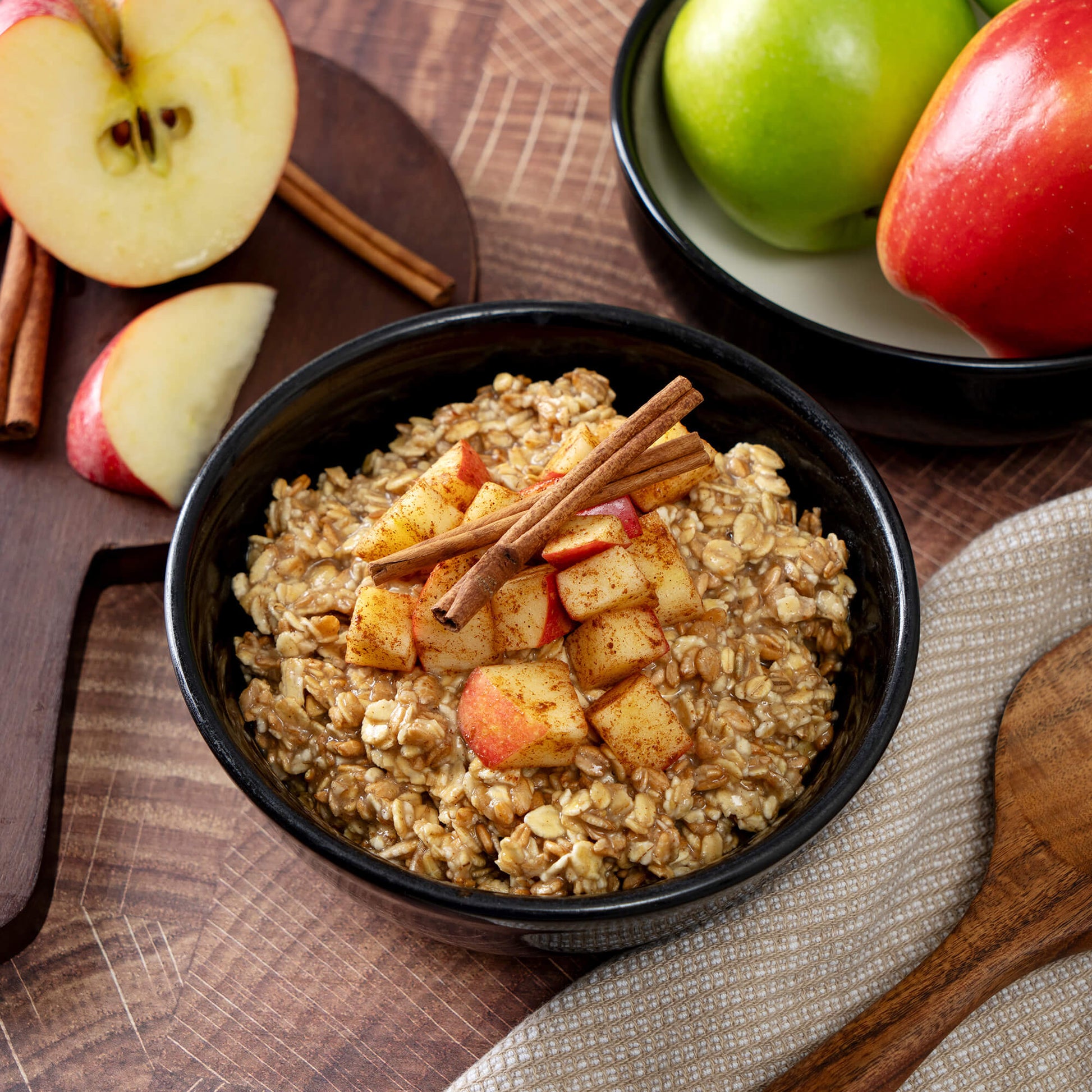 A bowl of oatmeal with chopped apples and cinnamon sits on a wooden table, with American Reserves 30 Day Emergency Food Supplys freeze-dried apples nearby, alongside whole apples and a cloth napkin—ideal for long-term storage.