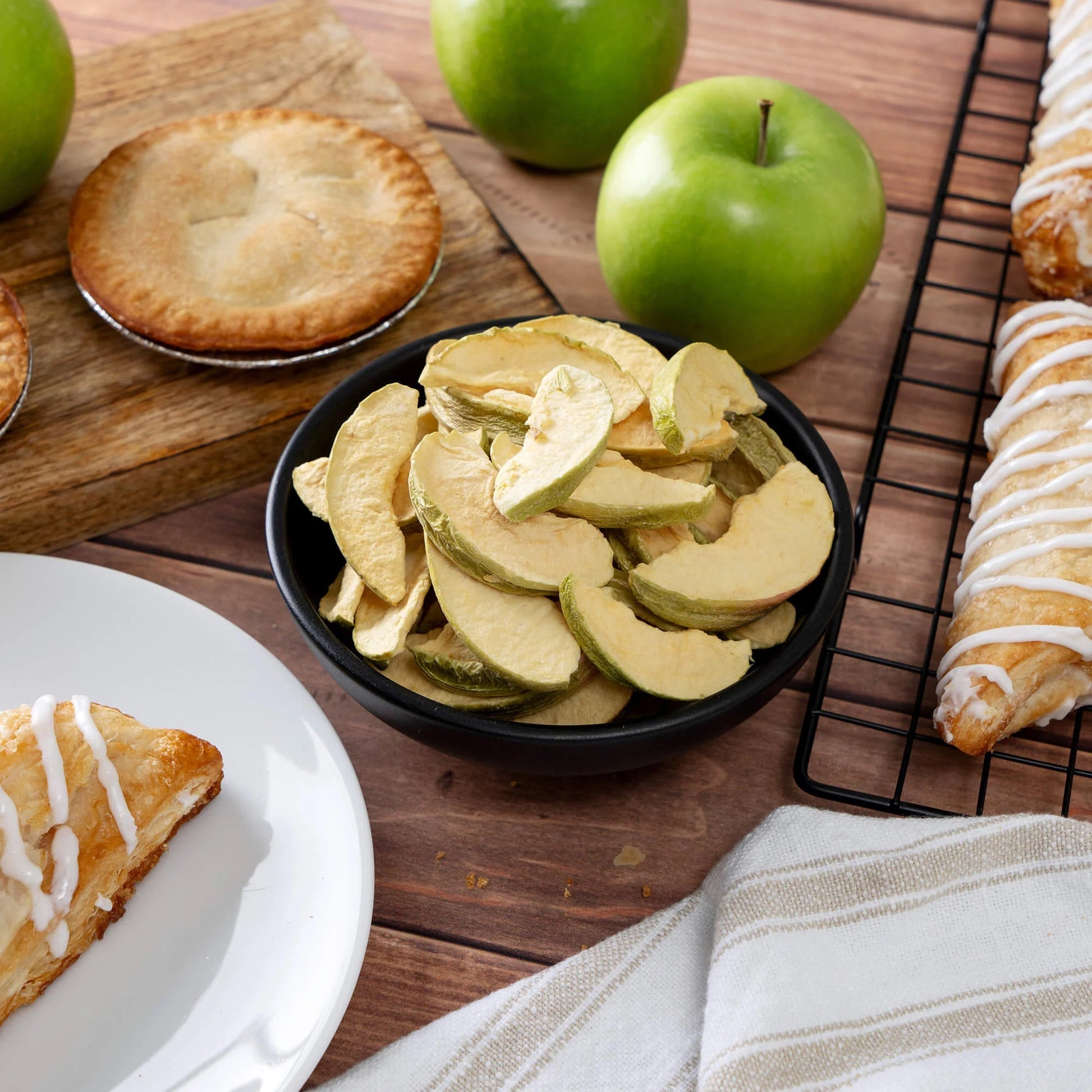 The 120 Serving Freeze Dried Fruit Bucket by American Reserves includes apple slices, perfect for a survival stash. Displayed with green apples, the bowl is accompanied by pies and a glazed pastry on a wooden board. A striped cloth napkin and white plate add charm to this essential emergency food item.