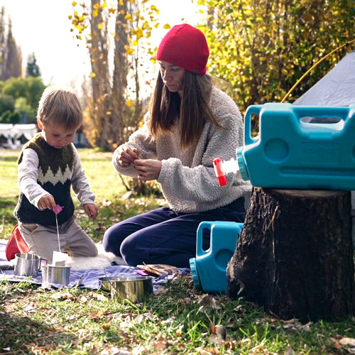 A woman and child sit outdoors by a tent, engaged in an activity. The child holds a skewer with white objects as AquaBrick 2 Pack Food and Water Storage Containers from American Reserves rest on a tree stump nearby. Tall trees provide a serene backdrop to their camping scene.