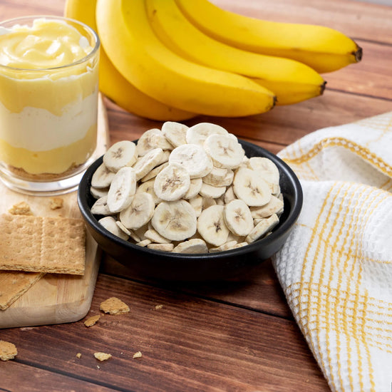On a wooden table, ripe bananas, a glass of layered banana pudding, and a bowl of banana chips are surrounded by graham crackers on a white and yellow checkered cloth—ideal comfort treats with a 25-year shelf-life, perfect for emergencies from American Reserves 30 Day Emergency Food Supply.