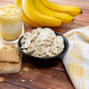 On a wooden table, ripe bananas, a glass of layered banana pudding, and a bowl of banana chips are surrounded by graham crackers on a white and yellow checkered cloth—ideal comfort treats with a 25-year shelf-life, perfect for emergencies from American Reserves 30 Day Emergency Food Supply.
