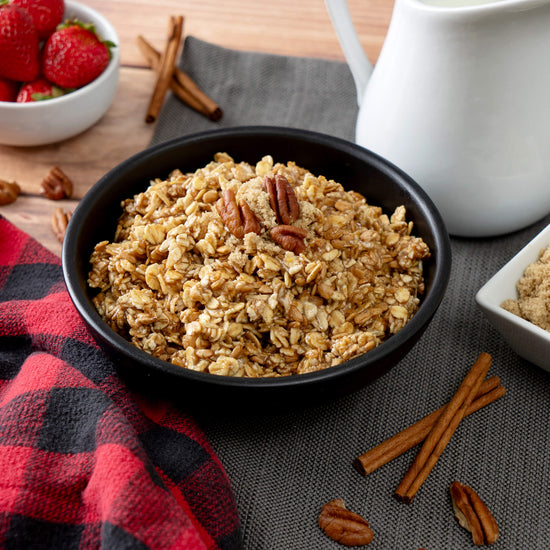 A bowl of granola with pecans sits on a dark cloth, with cinnamon sticks nearby, evoking the comfort of American Reserves 30 Day Emergency Food Supply. A jug, a small strawberry bowl, and a checkered red and black cloth enhance the scene.