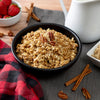 A bowl of granola with pecans sits on a dark cloth, with cinnamon sticks nearby, evoking the comfort of American Reserves 30 Day Emergency Food Supply. A jug, a small strawberry bowl, and a checkered red and black cloth enhance the scene.
