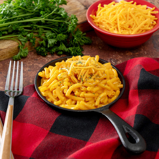 A skillet of macaroni and cheese, perfect for the American Reserves 30 Day Emergency Food Supply, is topped with shredded cheese and pepper on a red and black checkered cloth. A nearby fork complements the scene with a bowl of shredded cheese and fresh parsley in the background.