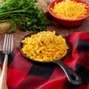A skillet of macaroni and cheese, perfect for the American Reserves 30 Day Emergency Food Supply, is topped with shredded cheese and pepper on a red and black checkered cloth. A nearby fork complements the scene with a bowl of shredded cheese and fresh parsley in the background.