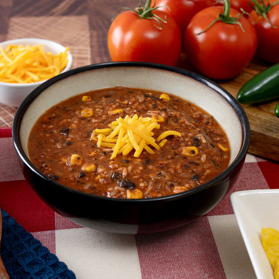 A bowl of chili topped with cheddar cheese, showcasing corn and beans, highlights American Reserves 30 Day Emergency Food Supply. Set on a red and white checkered cloth, fresh tomatoes, jalapeño, and extra cheese complete this long shelf-life meal.