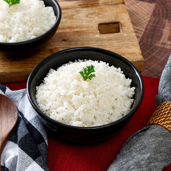 A black bowl with steamed white rice, topped with parsley, from American Reserves 30 Day Emergency Food Supply sits on a wooden surface with a red and white cloth nearby, another bowl partially visible in the background. Perfect for emergency preparedness.