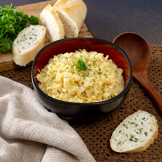 The 30 Day Emergency Food Supply by American Reserves offers a bowl of pasta with creamy sauce, garnished with green herbs, and paired with herb-topped bread. A wooden spoon, fresh parsley, and a small towel add charm, making it perfect to elevate any meal prep beyond typical emergency food.
