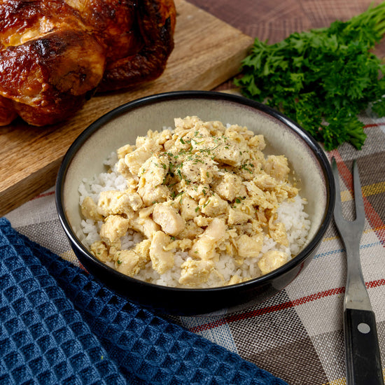 A bowl from the American Reserves 110 Serving Freeze Dried Meat & Sides Bucket features white rice with seasoned, cooked chicken pieces and herb garnish. In the background, a roasted chicken on a wooden board and fresh parsley create charm, while a fork rests on a checkered cloth nearby.