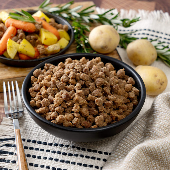 A bowl of cooked ground meat from the 110 Serving Freeze Dried Meat & Sides Bucket by American Reserves is on a table, surrounded by herbs and whole potatoes, emphasizing its long shelf life. In the background, cooked potatoes and carrots are in another bowl, with a fork resting on a cloth nearby.