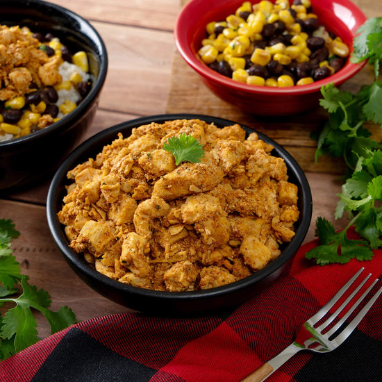 A black bowl filled with seasoned chicken from American Reserves 110 Serving Freeze Dried Meat & Sides Bucket is garnished with cilantro. Nearby, a red bowl holds corn and black beans. All bowls are set on a red and black cloth, ready to savor this feast.