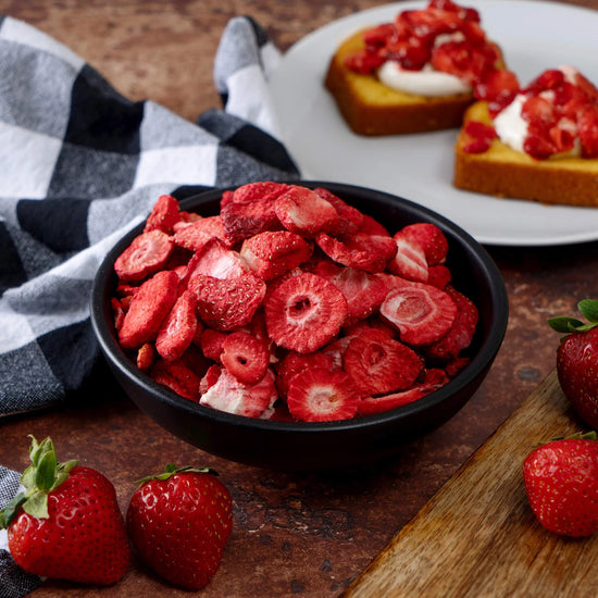 A black bowl filled with American Reserves freeze-dried strawberry slices from the 120 Serving Freeze Dried Fruit Bucket sits on a wooden table, alongside fresh strawberries. Toast topped with strawberries is on a plate next to a black-and-white checkered cloth in the background.