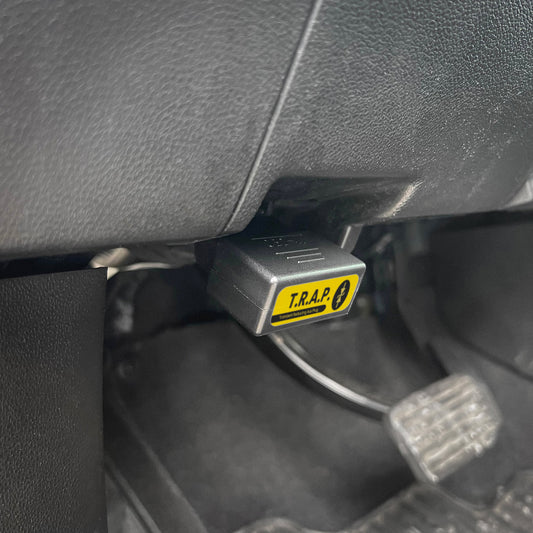 The T.R.A.P., a product of American Reserves, is securely connected to the cars OBD2 port above the pedals, providing EMP Vehicle Protection with transient voltage suppression for enhanced safety.