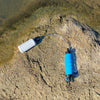 The RapidFlo Portable Water Filter by American Reserves, featuring a white inlet and blue handle, sits on a rock near water, promising safe hydration in the great outdoors.