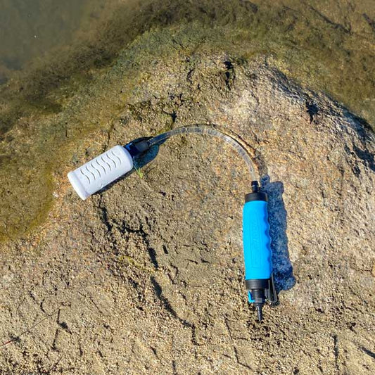 The RapidFlo Portable Water Filter by American Reserves, featuring a white inlet and blue handle, sits on a rock near water, promising safe hydration in the great outdoors.