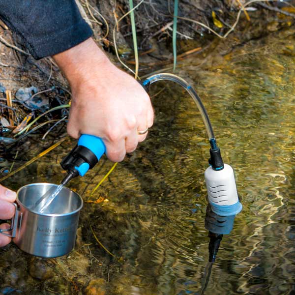 An individual uses the American Reserves RapidFlo Portable Water Filter to fill a metal cup from a natural water source, ideal for camping water filtration.