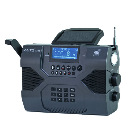 The Kaito Voyager Max KA900 by American Reserves is a black emergency radio featuring a digital display reading FM 106.8 MHz, buttons, knobs, a top solar panel, and an antenna. It includes a hand crank for emergencies and offers Bluetooth, MP3 playback, recording, weather alerts, and phone charging.