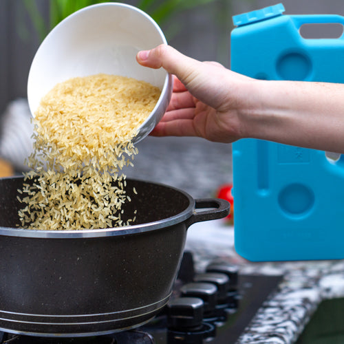 A hand pours uncooked rice into a black pot on the stove, with an American Reserves AquaBrick 2 Pack Food and Water Storage Container nearby, adding efficiency and style to the kitchen setup.