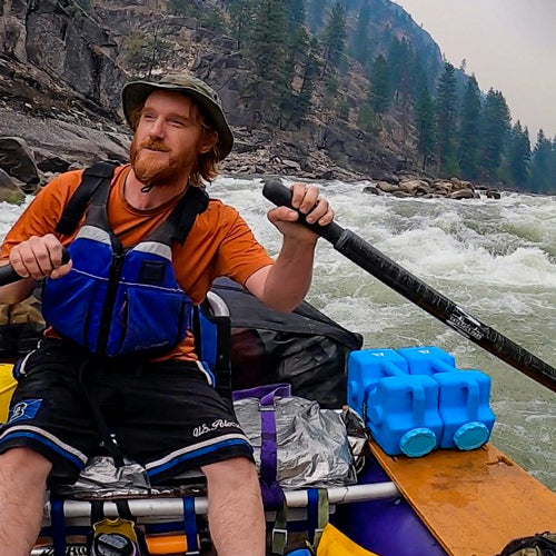 A person in an orange shirt and blue life jacket paddles a raft on a rocky river, their American Reserves AquaBrick 2 Pack Food and Water Storage Containers securely fastened for the adventure.