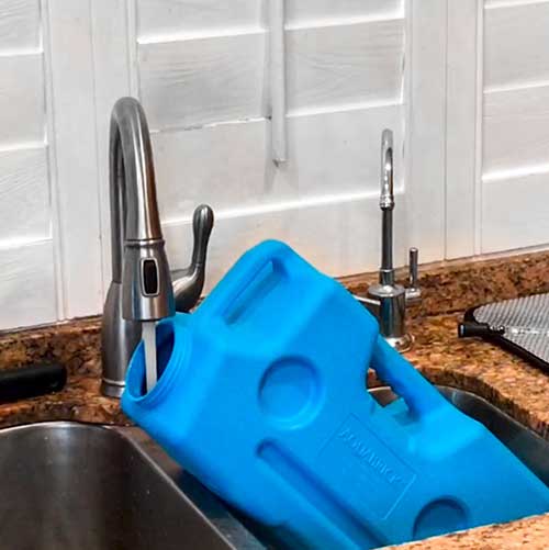 The AquaBrick 6 Pack, by American Reserves, is filled from a kitchen faucet in a sink set against a brown countertop. These large blue containers are perfect for water storage.
