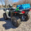 An ATV parked on gravel near water holds AquaBrick 6 Pack Food and Water Storage Containers from American Reserves, secured by a green strap on the rear rack. Bare trees line the backdrop, providing a picturesque scene for those equipped with water storage solutions.