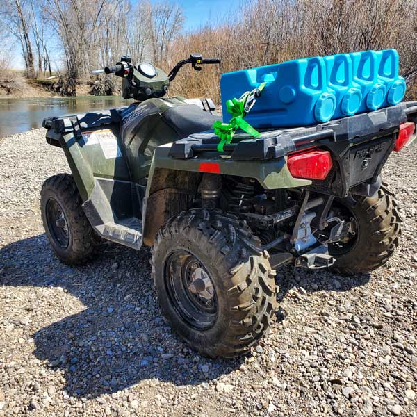 An ATV parked on gravel near water holds AquaBrick 6 Pack Food and Water Storage Containers from American Reserves, secured by a green strap on the rear rack. Bare trees line the backdrop, providing a picturesque scene for those equipped with water storage solutions.