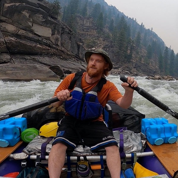 A bearded individual in a hat confidently navigates a rocky river raft amid lush trees, wearing an orange shirt and blue life vest, trusting the AquaBrick 6 Pack Food and Water Storage Containers by American Reserves for their water storage needs.