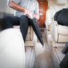 A person in a private jet reaches into a black bag on their lap for a book. The cream seats and exit sign are visible, as is the sleek design of the CORDURA® Berry Compliant Faraday Tablet Jacket by American Reserves, which provides signal blocking for uninterrupted focus.