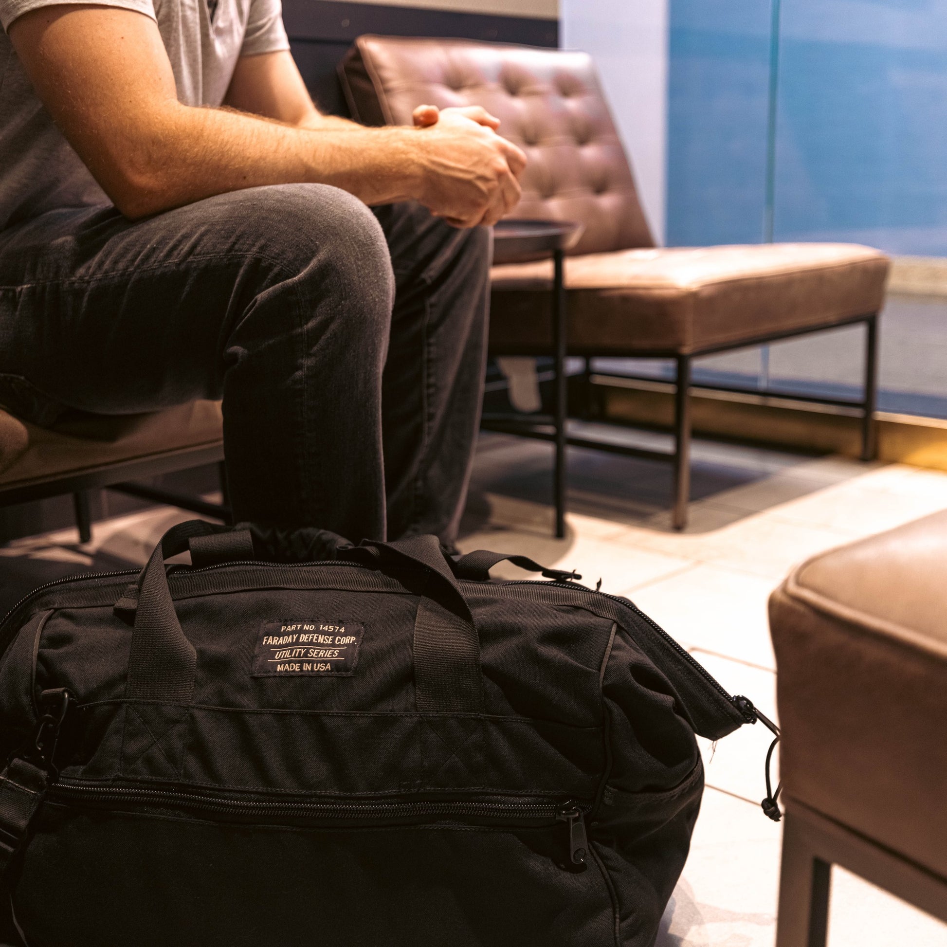 A person sits with hands clasped beside a rugged black American Reserves CORDURA® Utility Bag Large on the floor.
