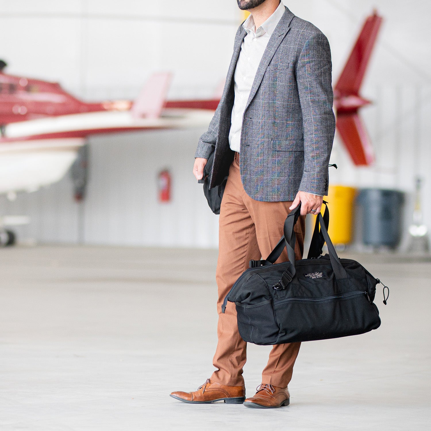 Amidst a vibrant red aircraft in an airplane hangar, someone wearing a blazer and brown pants carries the sleek American Reserves CORDURA® Utility Bag Medium.