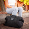 A person in a white top and jeans sits on a wooden ledge next to an American Reserves black CORDURA® Utility Bag Medium. A brick building and trees provide an urban backdrop.