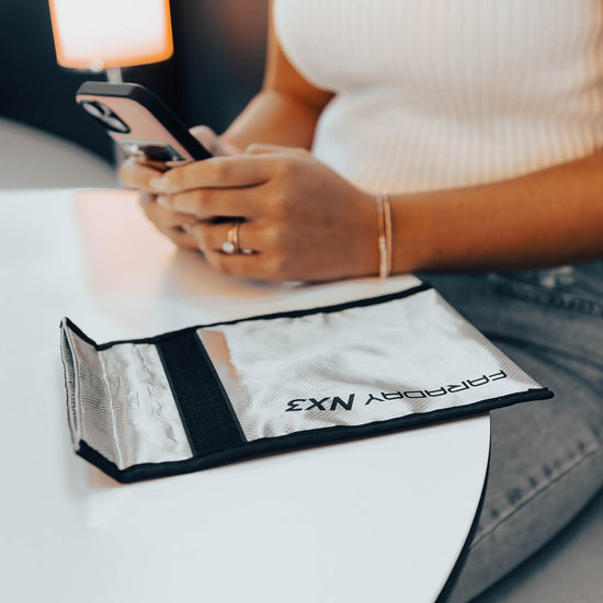 A person sits at a table using a smartphone, while an American Reserves Anti-Hacking and EMF Shielding Bag made from CYBER NC fabric is placed beside them.