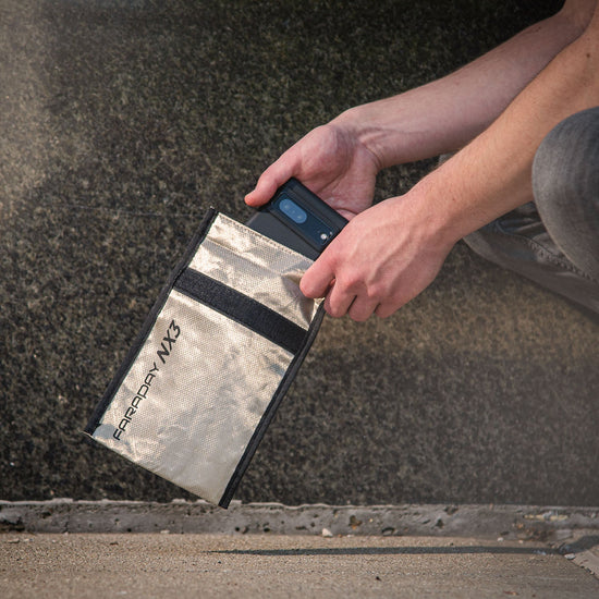 Someone carefully places a dark object into an Anti-Hacking and EMF Shielding Bag by American Reserves, its CYBER NC fabric gleaming subtly in the light near a textured surface.