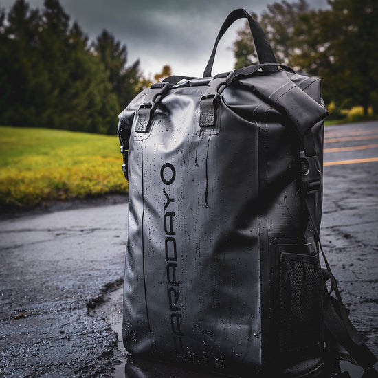 A Stealth Black Faraday Dry Bag Backpack from American Reserves, labeled FARADAY vertically, rests on a wet surface with trees in the background. It provides robust EMP protection to keep your devices secure while youre on the move.