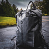 A Stealth Black Faraday Dry Bag Backpack from American Reserves, labeled FARADAY vertically, rests on a wet surface with trees in the background. It provides robust EMP protection to keep your devices secure while youre on the move.