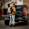 A person in a brown jacket with a backpack arranges items at the open tailgate of a pickup truck into an American Reserves Faraday Dry Bag Backpack, designed for EMP protection, in Stealth Black.