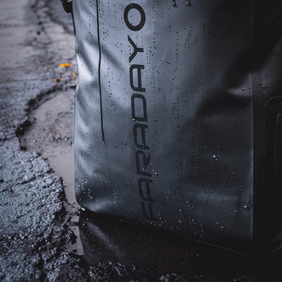 Close-up of a textured American Reserves Faraday Dry Bag Backpack in Stealth Black, featuring signal-blocking technology and FARADAY printed vertically, resting on rain-speckled asphalt.