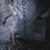 Close-up of a textured American Reserves Faraday Dry Bag Backpack in Stealth Black, featuring signal-blocking technology and FARADAY printed vertically, resting on rain-speckled asphalt.