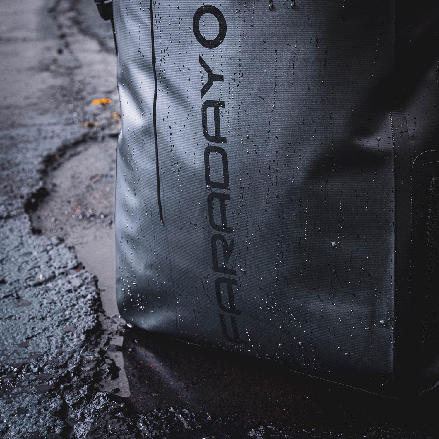 Close-up of a textured American Reserves Faraday Dry Bag Backpack in Stealth Black, featuring signal-blocking technology and FARADAY printed vertically, resting on rain-speckled asphalt.