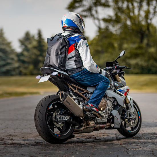 A motorcyclist, wearing a helmet and an American Reserves Stealth Black Faraday Dry Bag Backpack with EMP protection, sits on a sport bike facing away down a tree-lined road.