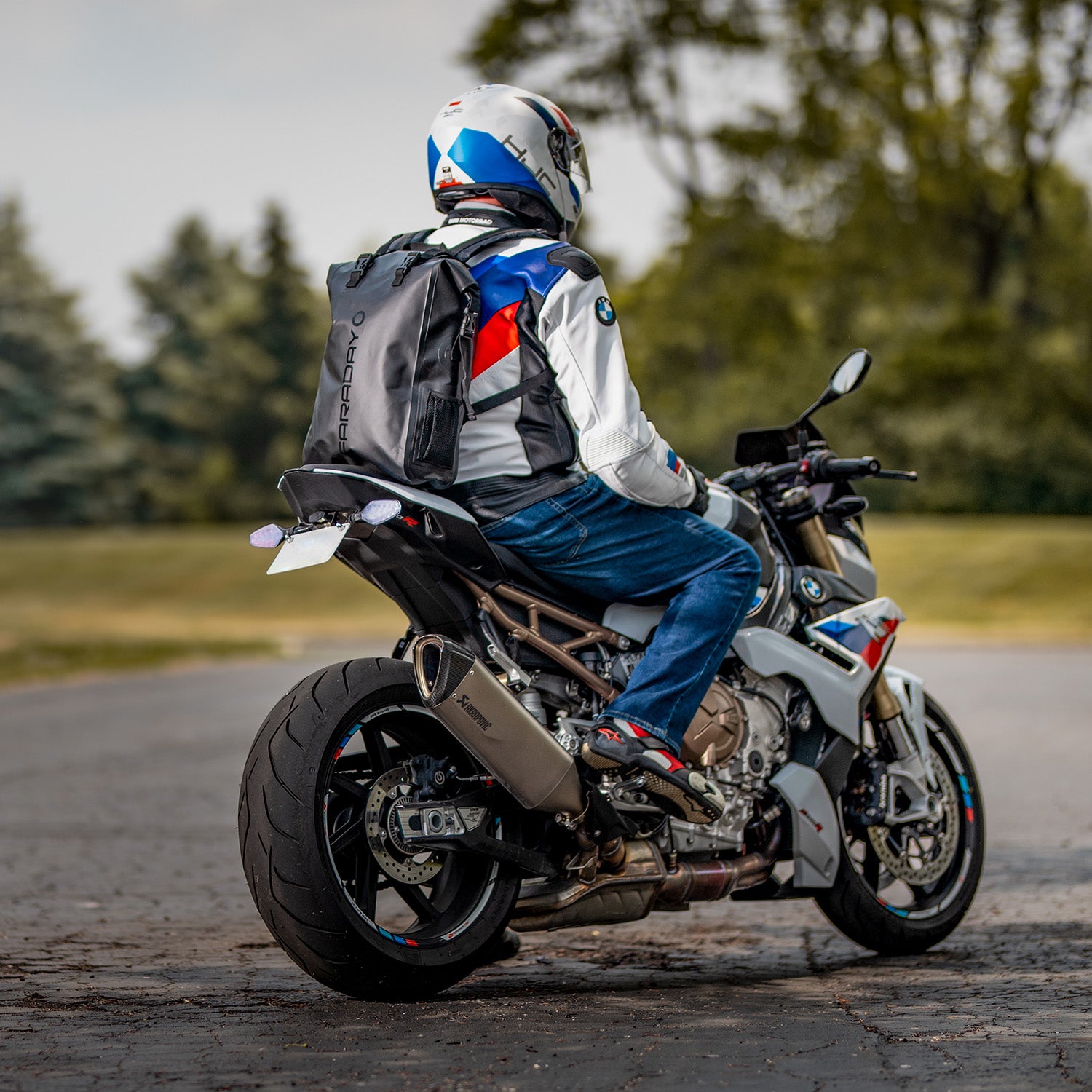 A motorcyclist, wearing a helmet and an American Reserves Stealth Black Faraday Dry Bag Backpack with EMP protection, sits on a sport bike facing away down a tree-lined road.