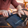 Someone in a maroon shirt is opening an American Reserves XL Forensic Faraday Jacket Kit, designed for signal blocking, on a metal outdoor table.