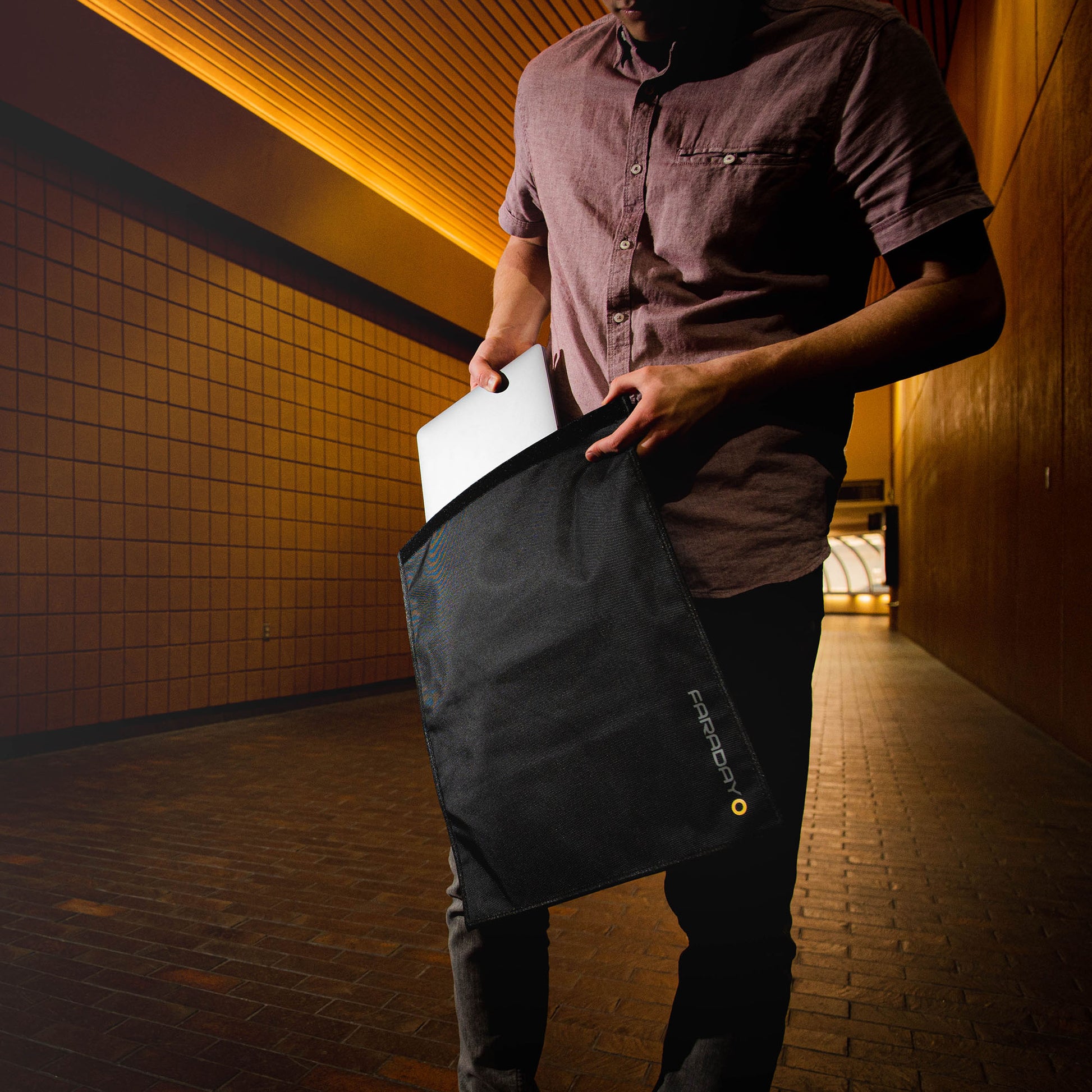 In a hallway, someone carefully slides their laptop into an XXL Forensic Faraday Laptop Jacket (14 x 16) by American Reserves, providing optimal device shielding.