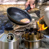 A person is cooking meat in a frying pan over a portable camping stove with a lively flame. Nearby, the American Reserves Kelly Kettle® Ultimate Trekker Camping Kit rests on rocky ground beside the gently flowing stream.